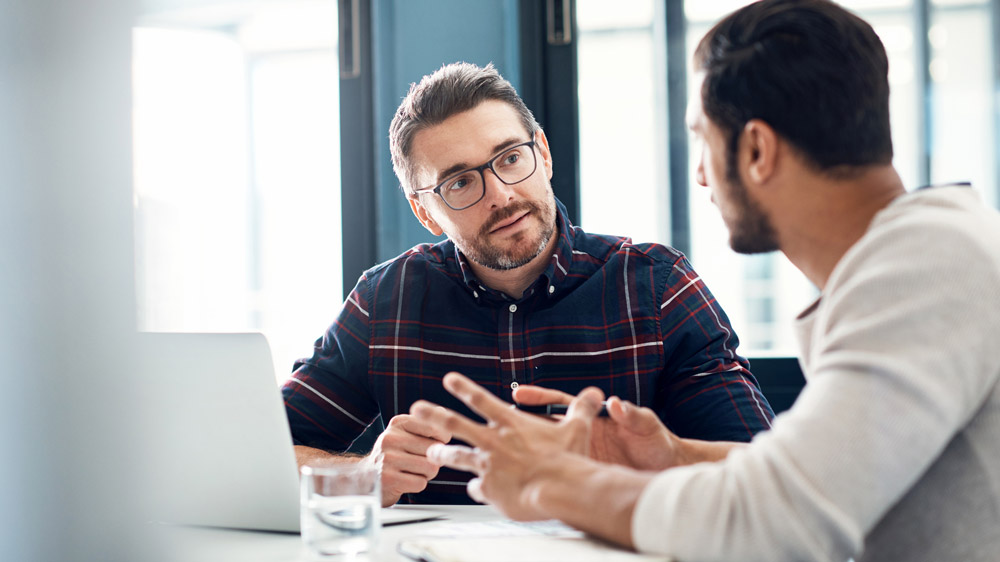 two men talking in office at desk with a computer