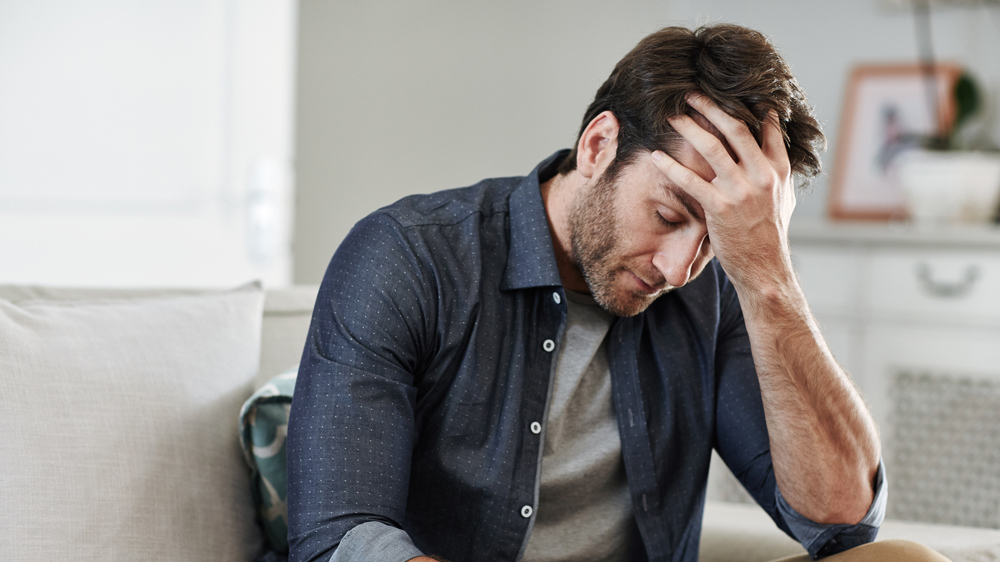 man sitting on couch with his head in hand, looking stressed and sad